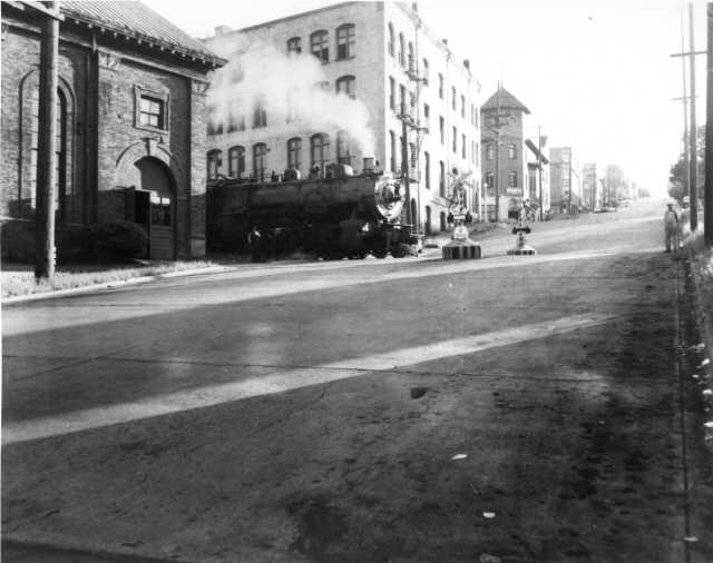 Undated image looking west up South 19th Street, Tacoma as a locomotive travels along the Prairie Line track. The Swiss Tavern is visible in background.   Tacoma Public Library, General Photograph Collection G44.1-130  