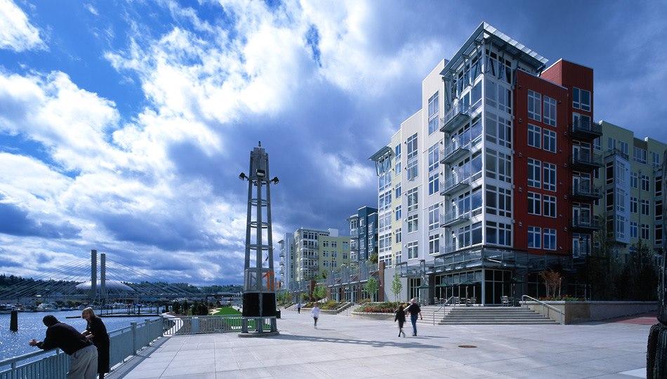 A color photo along the Thea Foss Waterway, showing pedestrians walking on the Thea Foss Esplanade, a broad walkway with landscaped features and condos surrounding it.