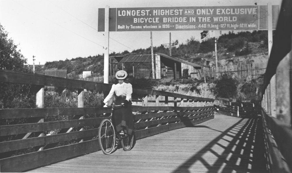 Image showing a woman crossing a wooden bicycle bridge in 1900, with a sign above reading &quot;Longest, Highest, and Only Exclusive Bicycle Bridge in the World.&quot; 